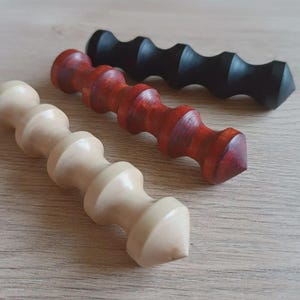 Puede incluir: Tres herramientas de masaje de madera en fila. Una es de color beige claro, otra marrón rojizo y la otra negra. Cada herramienta tiene una serie de secciones redondeadas y un extremo puntiagudo. Sobre una superficie de madera marrón claro.