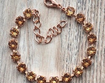 May include: A rose gold tone bracelet featuring a single row of round, faceted amber-colored crystals set in metal prongs. The bracelet includes a lobster claw clasp and an adjustable chain link extension for a secure and customizable fit.