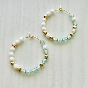 May include: A pair of gold hoop earrings with white and light blue beads. The beads are arranged in a gradient pattern, with the white beads on the top half of the hoop and the light blue beads on the bottom half.
