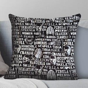 May include: Black throw pillow with a repeating pattern of human skull illustrations and anatomical terms like "skull," "ribs," and "vertebrae" in white. The pillow is square and rests on a gray surface.