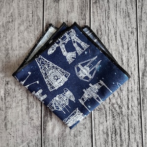 May include: A navy blue pocket square with a white outline of various Star Wars spaceships and vehicles. The pocket square is folded and has a black border.
