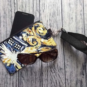 May include: A blue and yellow zippered pouch with a Starry Night-inspired design featuring a Dalek from Doctor Who. The pouch has a black strap and is lying on a wooden surface with a pair of sunglasses and a black smartphone.