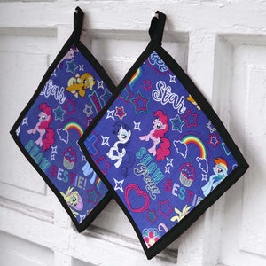 Könnte beinhalten: Zwei quadratische, blaue Topflappen mit schwarzem Rand und Aufhängeschlaufen. Die Topflappen zeigen ein farbenfrohes Muster aus Zeichentrickponys, Sternen, Regenbogen und den Worten "Show Bestie!" und "Star".