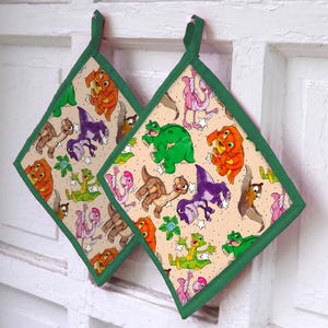 May include: Two square pot holders with a beige background and a repeating pattern of colorful cartoon dinosaurs. The pot holders have green trim and hanging loops. The dinosaurs are in shades of orange, green, purple, pink, and brown.