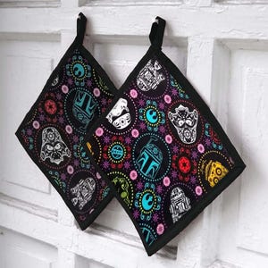 May include: Two black square pot holders with a Star Wars-themed design. The design features various characters and symbols in white, teal, pink, and yellow, set against a black background. Each pot holder has a black loop for hanging.