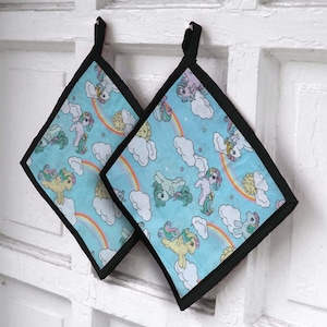 May include: Two square pot holders with a light blue background featuring cartoon ponies, rainbows, clouds, and stars. The pot holders have a black border and a black loop for hanging.