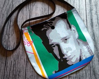 Könnte beinhalten: Eine kleine, runde Umhängetasche mit schwarzem Riemen. Die Tasche zeigt einen grafischen Druck eines Männergesichts in Grau-, Grün- und Blautönen. Das Design umfasst farbige Streifen und eine handschriftliche Signatur.