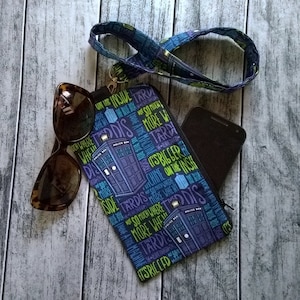 Peut inclure: Une pochette zippée bleue et violette pour téléphone avec une lanière, des lunettes de soleil et un smartphone. L'étui présente un motif répété du TARDIS et du texte. Les lunettes de soleil ont une monture écaille de tortue.