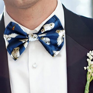 May include: A navy blue bow tie with a white owl pattern. The bow tie is tied around a white shirt collar and a dark blue suit jacket. A small bouquet of white flowers is pinned to the jacket.