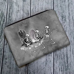 May include: A gray zippered pouch with a design of three skeletons dancing. The skeletons are white and have black outlines. The background is a gray watercolor wash.