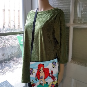 Puede incluir: Un peque&ntilde;o bolso cruzado blanco con una colorida ilustraci&oacute;n de Ariel de La Sirenita. El bolso tiene una correa negra y lo lleva un maniqu&iacute;. La ilustraci&oacute;n incluye a Ariel, Flounder, Sebasti&aacute;n y otras criaturas marinas.