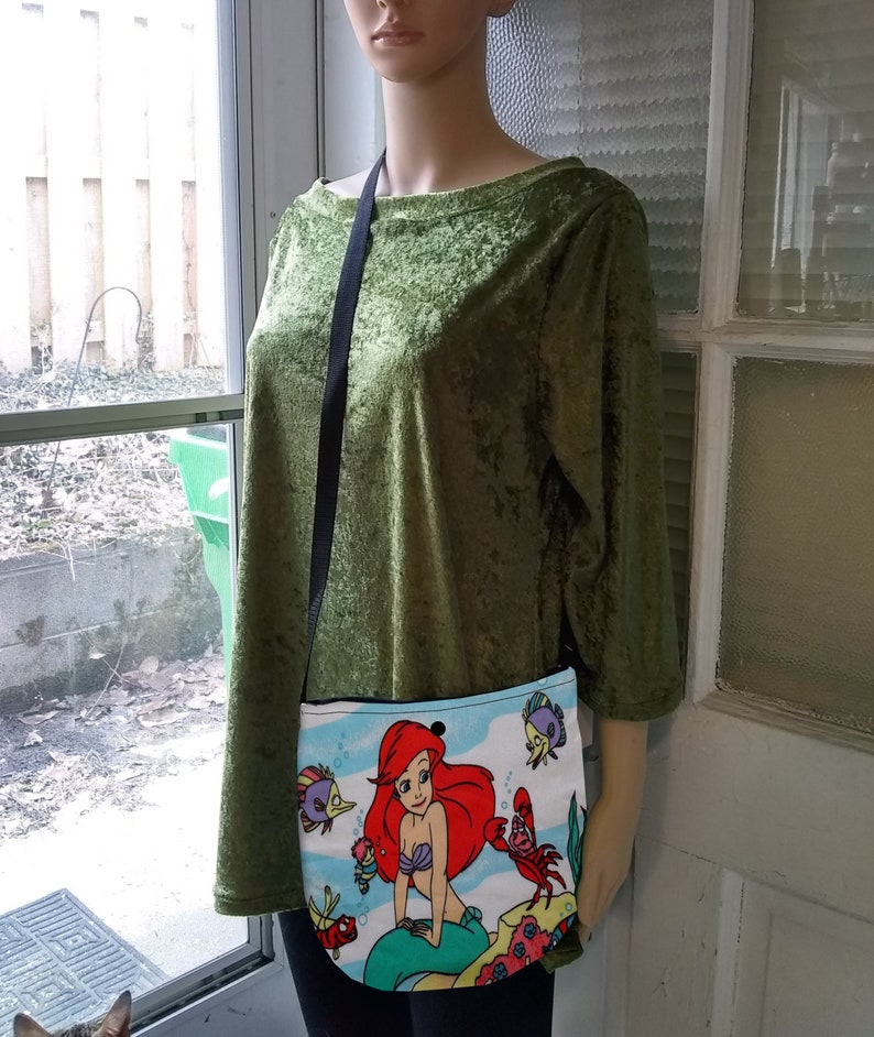 Puede incluir: Un peque&ntilde;o bolso cruzado blanco con una colorida ilustraci&oacute;n de Ariel de La Sirenita. El bolso tiene una correa negra y lo lleva un maniqu&iacute;. La ilustraci&oacute;n incluye a Ariel, Flounder, Sebasti&aacute;n y otras criaturas marinas.