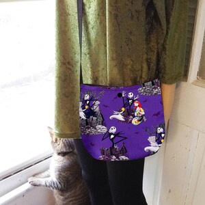 Puede incluir: Un bolso bandolera morado con un patr&oacute;n repetido de personajes y escenas de la pel&iacute;cula "Pesadilla antes de Navidad". El bolso tiene una correa negra y una forma curva. El fondo incluye im&aacute;genes de una casa embrujada y murci&eacute;lagos.
