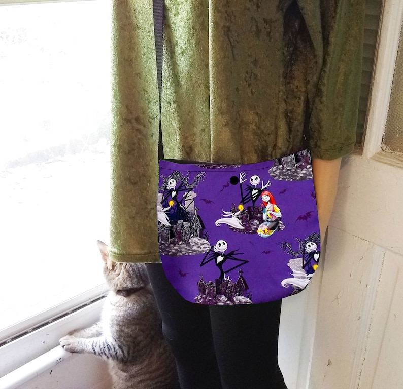 Puede incluir: Un bolso bandolera morado con un patr&oacute;n repetido de personajes y escenas de la pel&iacute;cula "Pesadilla antes de Navidad". El bolso tiene una correa negra y una forma curva. El fondo incluye im&aacute;genes de una casa embrujada y murci&eacute;lagos.