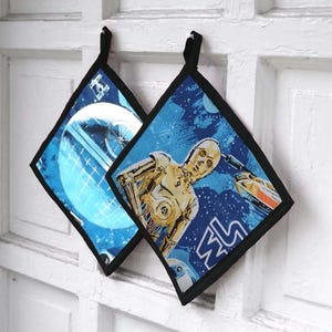 Può includere: Due presine a forma di diamante con bordi neri e anelli per appenderle. Una presenta un design della Morte Nera, l'altra C-3PO e un'astronave su uno sfondo blu stellato. Le presine sono in tessuto.