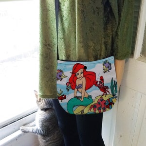Puede incluir: Un peque&ntilde;o bolso cruzado azul claro con una colorida ilustraci&oacute;n de Ariel de La Sirenita. El bolso tiene una correa negra. El dise&ntilde;o incluye a Ariel, peces y otras criaturas marinas.