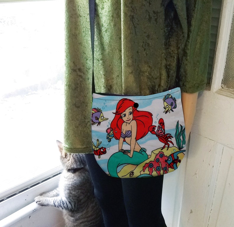 Puede incluir: Un peque&ntilde;o bolso cruzado azul claro con una colorida ilustraci&oacute;n de Ariel de La Sirenita. El bolso tiene una correa negra. El dise&ntilde;o incluye a Ariel, peces y otras criaturas marinas.