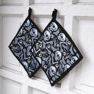 May include: Two black oven mitts with a skull and crossbones pattern. The mitts are made of a black fabric with a white skull and crossbones design. The mitts are hanging on a white wall.
