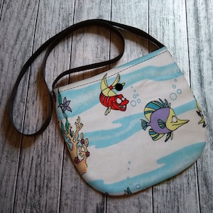 Puede incluir: Bolso cruzado blanco y azul con estampado de peces de dibujos animados. El bolso tiene una correa negra y un cierre de bot&oacute;n. El dise&ntilde;o incluye peces de dibujos animados, burbujas y coral.