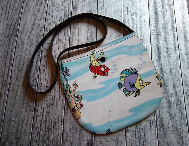 Puede incluir: Bolso cruzado blanco y azul con estampado de peces de dibujos animados. El bolso tiene una correa negra y un cierre de bot&oacute;n. El dise&ntilde;o incluye peces de dibujos animados, burbujas y coral.