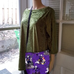 Puede incluir: Un bolso bandolera morado con un estampado de personajes de Pesadilla antes de Navidad. El bolso tiene una correa negra y se muestra en un maniqu&iacute; con una blusa de terciopelo verde.