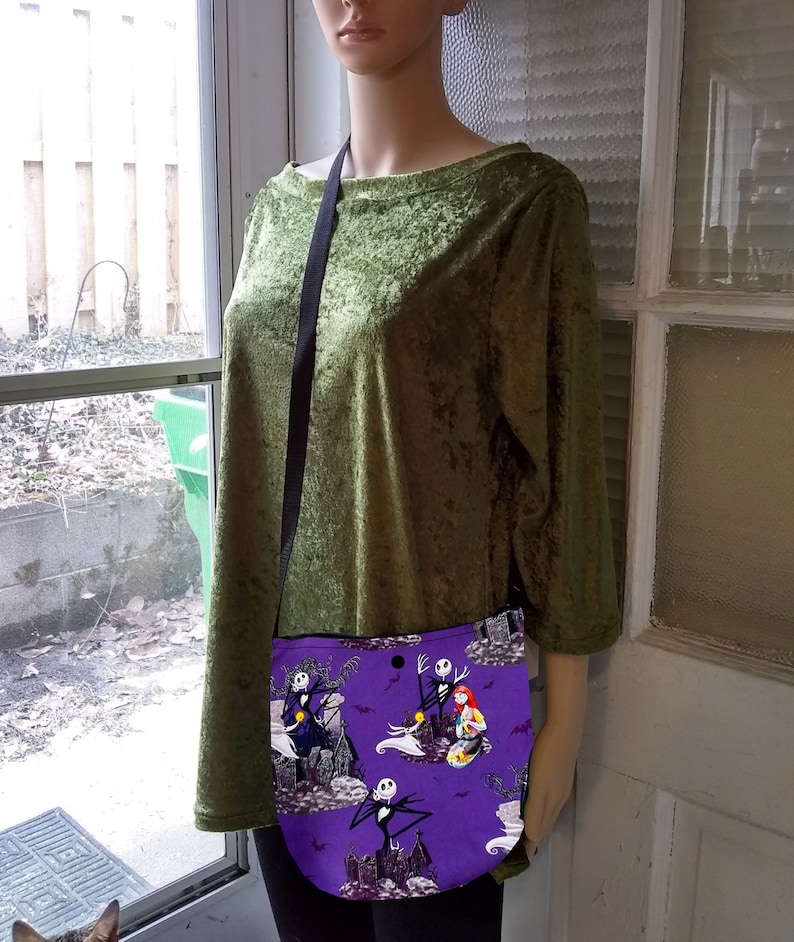 Puede incluir: Un bolso bandolera morado con un estampado de personajes de Pesadilla antes de Navidad. El bolso tiene una correa negra y se muestra en un maniqu&iacute; con una blusa de terciopelo verde.