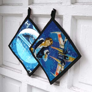 May include: Two square pot holders with black trim and hanging loops. One features a Death Star design, the other a scene with a character and X-wing fighters, all on a blue starry background. The pot holders are likely made of fabric.