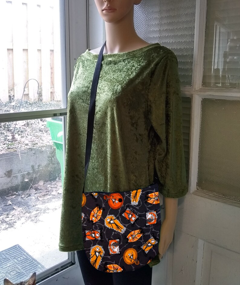 K&ouml;nnte beinhalten: Eine schwarze Umh&auml;ngetasche mit einem Halloween-Motiv, das orangefarbene und wei&szlig;e Jack-Skellington-Figuren auf schwarzem Hintergrund zeigt.