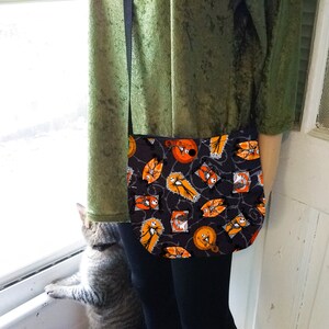 K&ouml;nnte beinhalten: Eine schwarze Umh&auml;ngetasche mit einem Halloween-Motiv, das Jack Skellington und Sally aus Nightmare Before Christmas zeigt. Der Druck ist orange, schwarz und wei&szlig;. Die Tasche hat einen schwarzen verstellbaren Riemen.