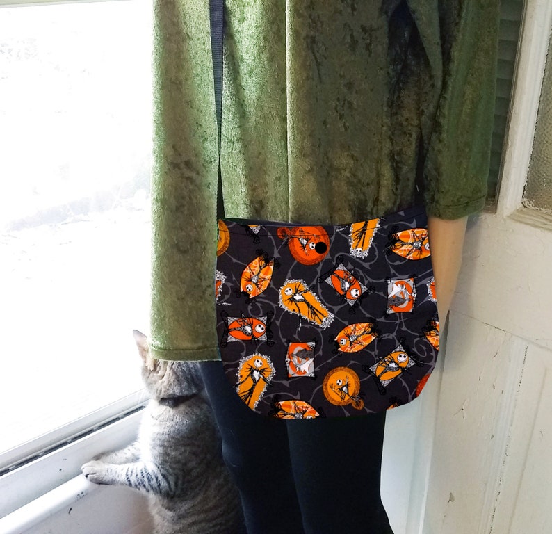 K&ouml;nnte beinhalten: Eine schwarze Umh&auml;ngetasche mit einem Halloween-Motiv, das Jack Skellington und Sally aus Nightmare Before Christmas zeigt. Der Druck ist orange, schwarz und wei&szlig;. Die Tasche hat einen schwarzen verstellbaren Riemen.