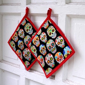 May include: Two black and white kitchen pot holders with a red trim. The pot holders feature a colorful sugar skull design.