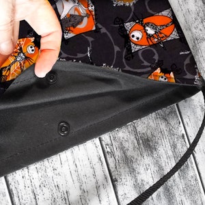 K&ouml;nnte beinhalten: Schwarze Stofftasche mit einem schwarzen und orangefarbenen Druck, der einen Comic-Charakter zeigt. Die Tasche hat einen schwarzen Kordelzug und zwei Druckkn&ouml;pfe.