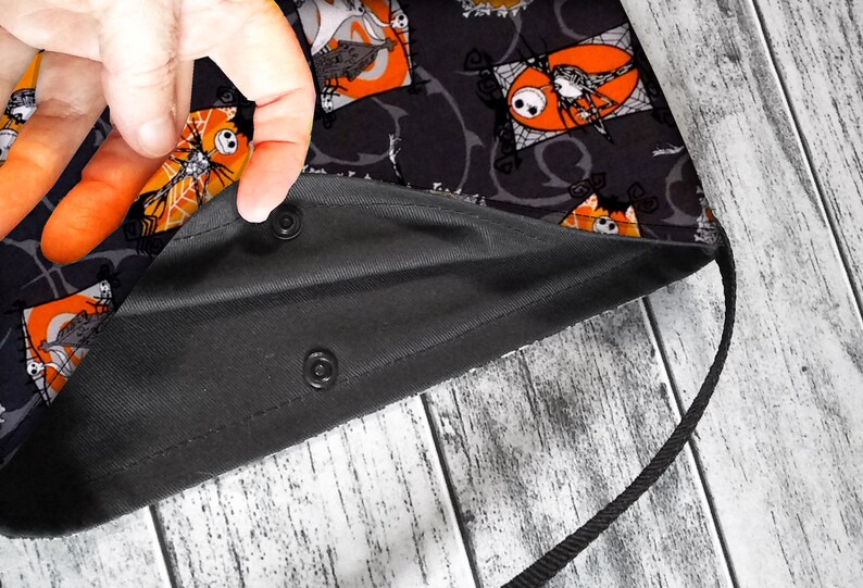 K&ouml;nnte beinhalten: Schwarze Stofftasche mit einem schwarzen und orangefarbenen Druck, der einen Comic-Charakter zeigt. Die Tasche hat einen schwarzen Kordelzug und zwei Druckkn&ouml;pfe.