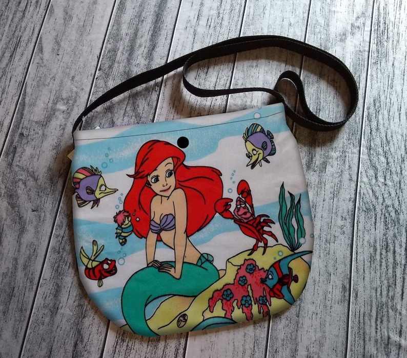 Puede incluir: Bolso de hombro de tela azul claro y blanco con una ilustraci&oacute;n de dibujos animados de Ariel la sirena, peces y un cangrejo rojo. El bolso tiene una correa negra y un cierre de bot&oacute;n a presi&oacute;n negro.