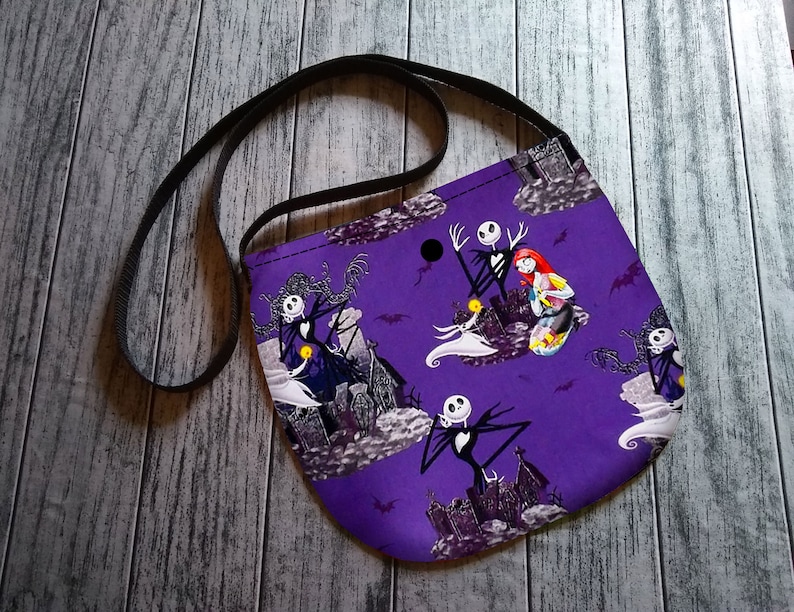 Puede incluir: Un bolso bandolera morado con un estampado repetido de personajes de Pesadilla antes de Navidad, incluyendo a Jack Skellington y Sally. El bolso tiene una correa negra y est&aacute; hecho de tela. El fondo incluye im&aacute;genes de una casa encantada y murci&eacute;lagos.