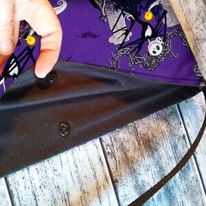 Puede incluir: Un bolso tote morado con interior negro y correa negra. El bolso presenta un estampado tem&aacute;tico de Halloween con personajes y motivos en el exterior. El bolso se asegura con cierres de presi&oacute;n negros.