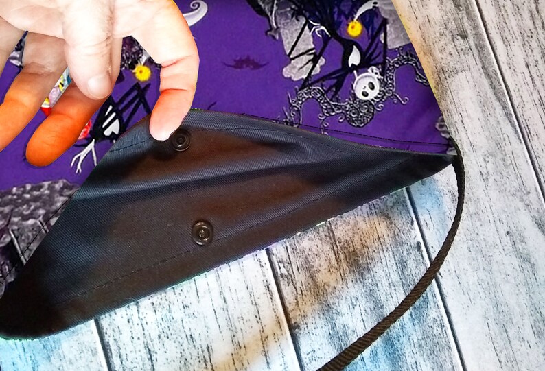 Puede incluir: Un bolso tote morado con interior negro y correa negra. El bolso presenta un estampado tem&aacute;tico de Halloween con personajes y motivos en el exterior. El bolso se asegura con cierres de presi&oacute;n negros.