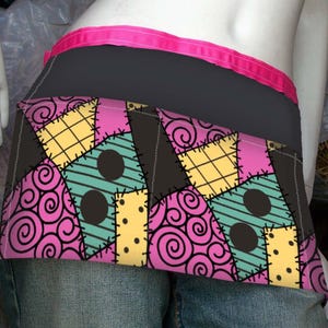 Könnte beinhalten: Schwarze und rosa Schürze mit Patchwork-Design. Die Schürze hat einen schwarzen Bund mit rosa Besatz. Das Patchwork-Design umfasst rosa Wirbel, gelbe Quadrate und türkisfarbene Dreiecke mit schwarzen Kreisen.