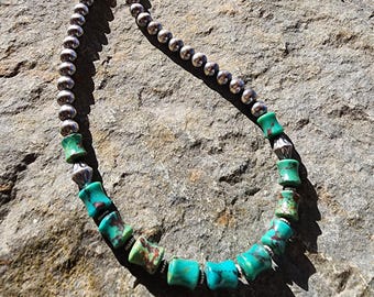 May include: A necklace featuring silver beads and turquoise elements. The necklace combines small, round silver beads with turquoise barrel-shaped beads and silver spacers. The jewellery is displayed on a textured stone surface.