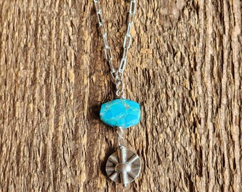 May include: A silver necklace with a turquoise stone and a sunburst pendant. The chain is a link style, and the pendant is round with a textured design. The necklace is displayed on a wooden surface.