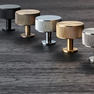 May include: Five cabinet knobs in a row, each with a textured, cylindrical head and stem. Finishes include black, brass, silver, and gold. The knobs are displayed on a dark wooden surface. The knobs are suitable for furniture.