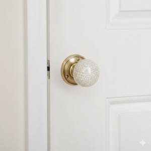 May include: A brass door knob with a round, crackle-glazed ceramic handle. The knob is mounted on a white door with a recessed panel. The door is slightly ajar, revealing the door frame and latch.