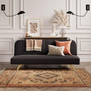 May include: A dark grey sofa with gold-coloured legs, decorated with cushions and a fringed throw. The sofa is placed on a patterned rug, with framed artwork and decorative items in the background. Two black wall sconces are mounted on the wall.