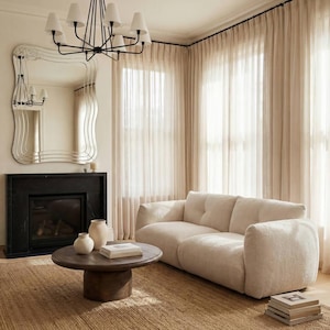 May include: A neutral-toned living room with a cream sofa, dark wood coffee table, and a black fireplace. A large, wavy-edged mirror is above the fireplace. Sheer curtains filter the light, and a black chandelier hangs from the ceiling.