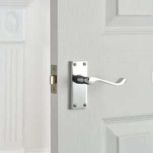 May include: A polished chrome door handle and backplate are fixed to a white panelled door. The handle features a curved lever design. The door is ajar, showing a white wall and trim.