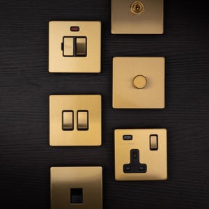 May include: A selection of gold-coloured electrical switch plates and sockets. The plates feature different designs, including toggle switches, a dimmer, a USB outlet, and a single socket. The background is dark wood grain.