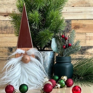 May include: A wooden gnome figurine with a white beard and a red hat. The gnome is surrounded by small red and green Christmas ornaments and a small potted evergreen tree.