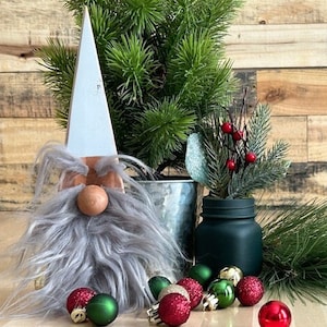 May include: A gray and white wooden gnome with a gray fur beard and a white pointed hat. The gnome is sitting on a wooden surface with green and red Christmas ornaments.