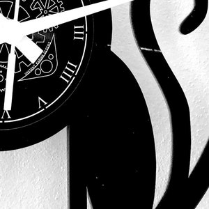 May include: Black and white wall clock with Roman numerals and clock hands. The clock face features the text "Instant Karma Clocks" and a gear design. The clock is mounted on a textured white wall.