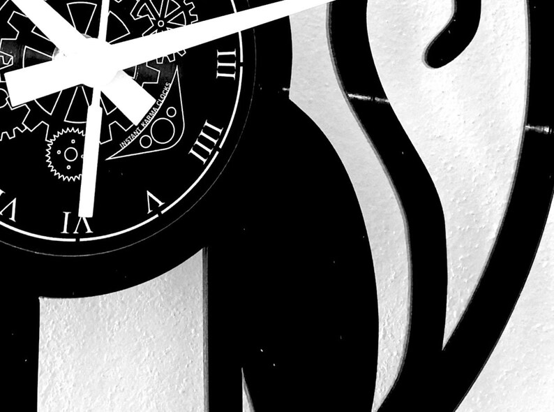 May include: Black and white wall clock with Roman numerals and clock hands. The clock face features the text "Instant Karma Clocks" and a gear design. The clock is mounted on a textured white wall.